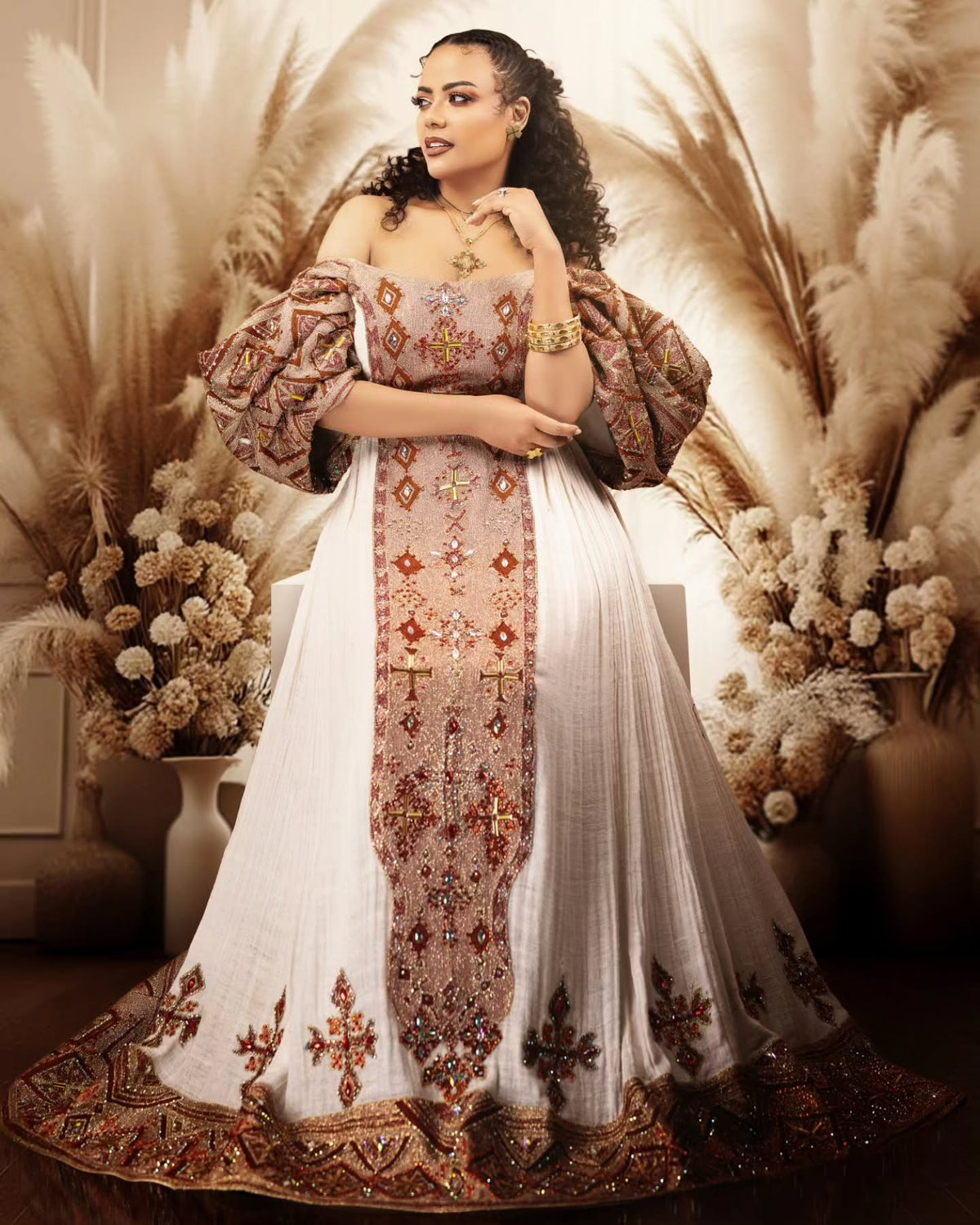 A woman models a traditional off-the-shoulder white and beige Habesha Wedding Dress with intricate gold, brown, and red embroidery, styled with a matching embroidered border and voluminous sleeves, set against a warm bohemian backdrop of pampas grass.