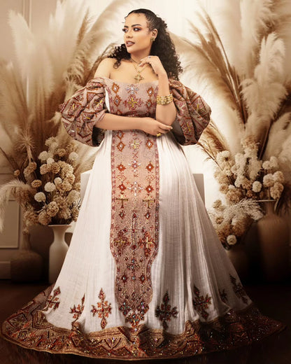 A woman models a traditional off-the-shoulder white and beige Habesha Wedding Dress with intricate gold, brown, and red embroidery, styled with a matching embroidered border and voluminous sleeves, set against a warm bohemian backdrop of pampas grass.