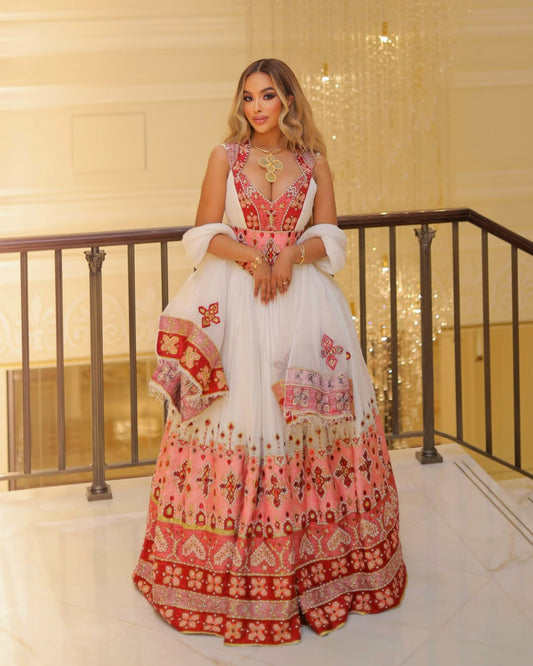 A woman stands indoors wearing a traditional embroidered habesha Wedding dress with a white upper section, pink and red heavily patterned lower skirt, and a matching sheer white dupatta draped over her arms. The setting features a warm-toned interior staircase with a black railing.