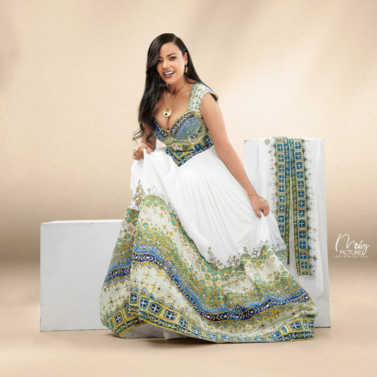 A woman models a white sleeveless habesha Wedding dress with ornate blue and green embroidered borders, standing beside a matching draped fabric panel on a beige studio background.