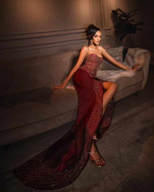 A woman models a strapless floor-length Habesha Wedding Dress in deep burgundy with intricate gold beading and sequins, posed on a gray sofa in a dimly lit room.