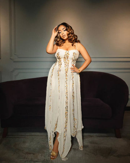 A woman models a strapless white high-low Gold Habesha Wedding Dress  with gold floral embroidery, standing in front of a dark purple velvet sofa in a dimly lit room.