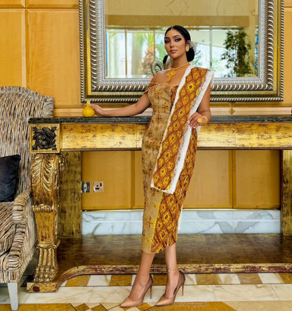 A woman models a sleeveless, off-the-shoulder midi habesha Wedding dress with a gold and brown geometric pattern and a white lace-trimmed neckline, standing in an ornate, luxurious room with gold paneling and a console table.