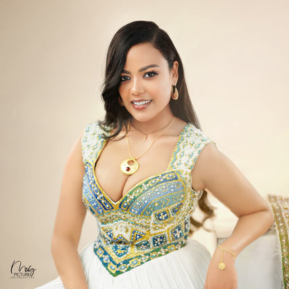 A woman wearing a heavily embellished green and gold sleeveless top with a deep V-neck, paired with a white habesha Wedding dress, posing against a warm beige background.