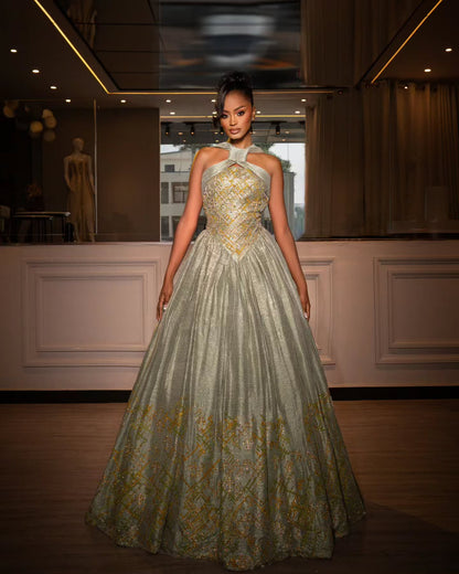 A woman models a strapless ball Habesha Wedding Dress with a halter-style neckline, featuring a fitted bodice with gold embroidery, a voluminous light gray-green skirt with gold floral patterns, and a floor-length train in an indoor boutique setting.