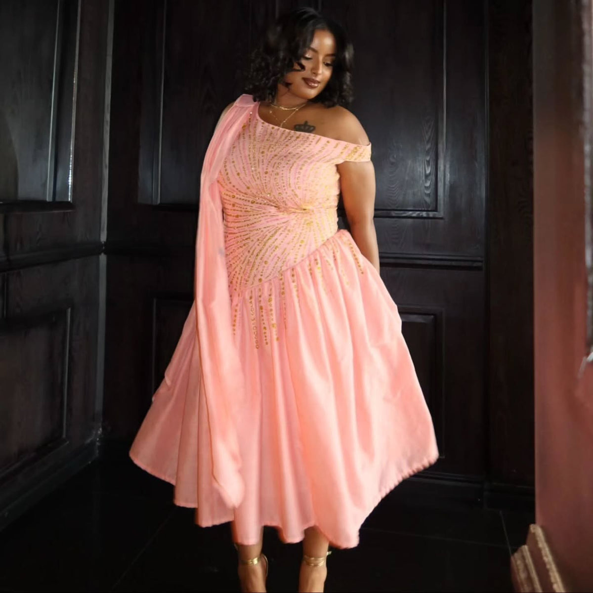 A woman wearing a peach-pink one-shoulder pink Habesha Wedding Dress with gold beaded embellishments and a matching draped scarf, standing in front of dark wooden paneling.