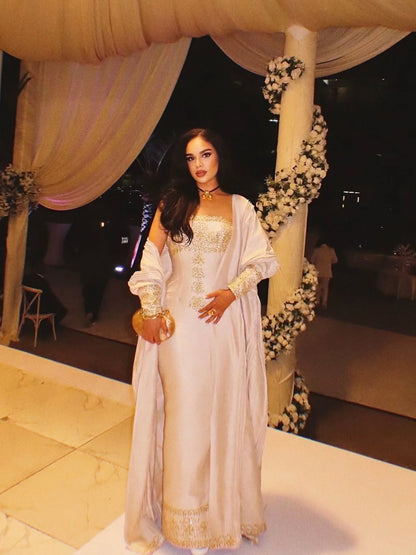 A woman stands in a decorated event space wearing a long, light beige or off-white traditional Gold Habesha Wedding Dress  with gold embroidery and a matching sheer draped overlay. The setting features draped cream fabric, floral garlands on the columns, and a dark nighttime background.