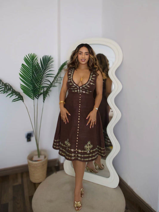 A woman models a brown brown Habesha wedding dress with gold embroidery and button details, standing in front of a white wavy mirror in a room with a potted plant.
