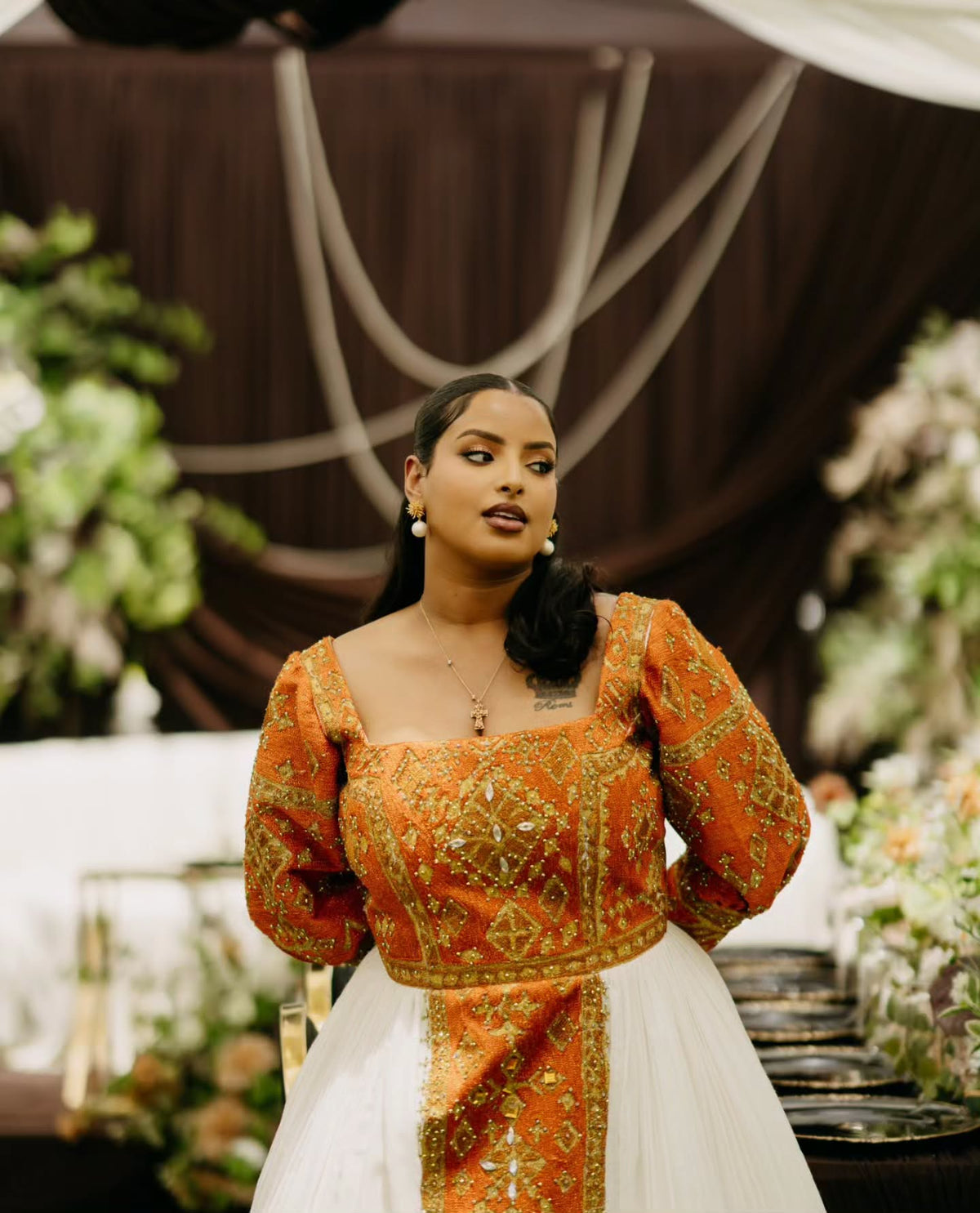 A woman wearing a traditional Habesha Wedding Dress  with a vibrant orange and gold embroidered bodice, puffed sleeves, and a white tulle skirt, standing in an elegant event setting with floral arrangements and draped brown fabric in the background.