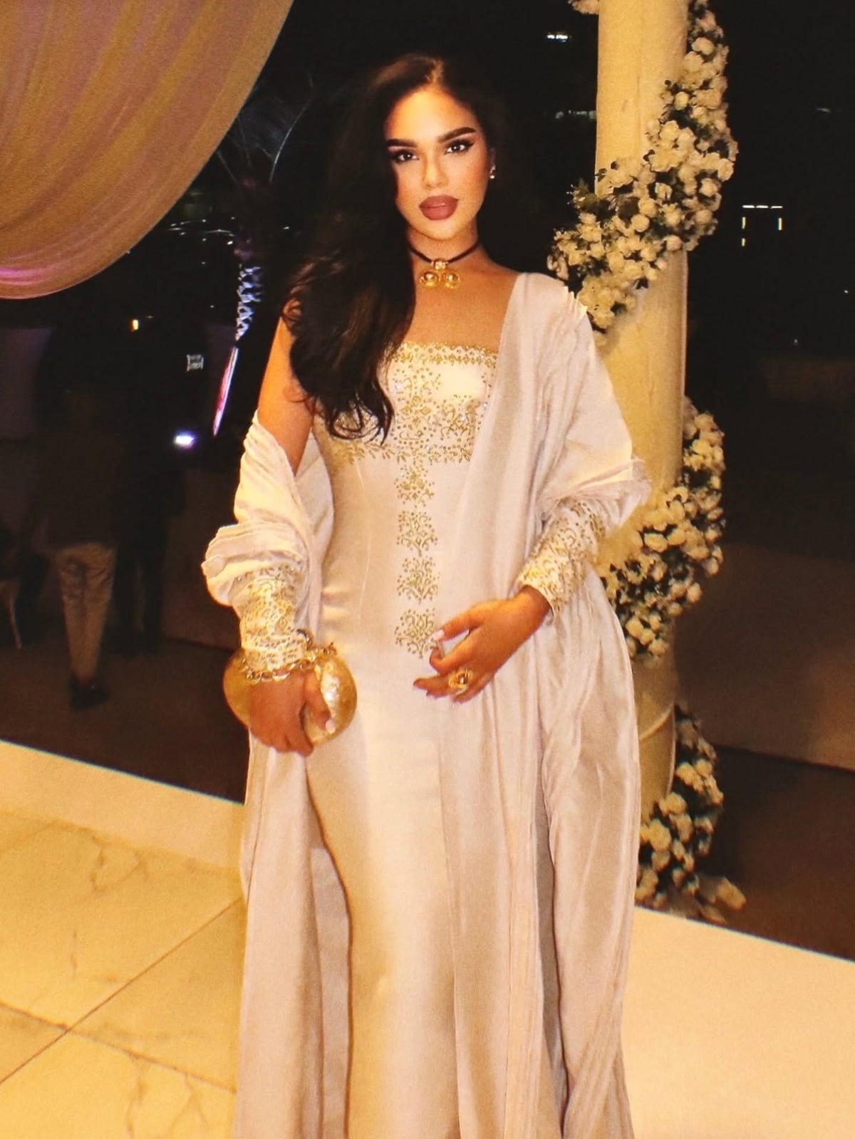 A woman wearing a long white traditional Gold Habesha Wedding Dress  with gold embroidery and a matching draped outer layer, standing in an indoor event setting with decorative columns and floral arrangements.