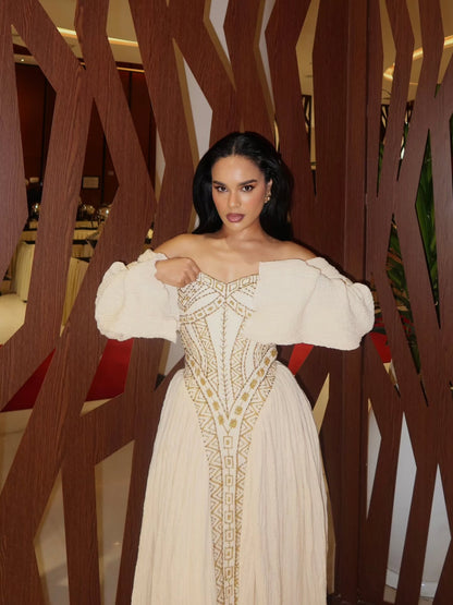 Woman in a white off-shoulder Habesha Wedding Dress with gold embroidery standing in front of a decorative wooden screen.