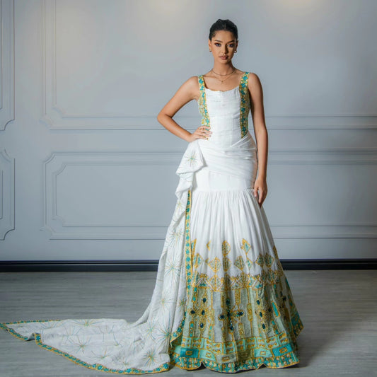 A woman models a white sleeveless Habesha Wedding Dress with green and gold embroidered details, standing against a light gray paneled wall.