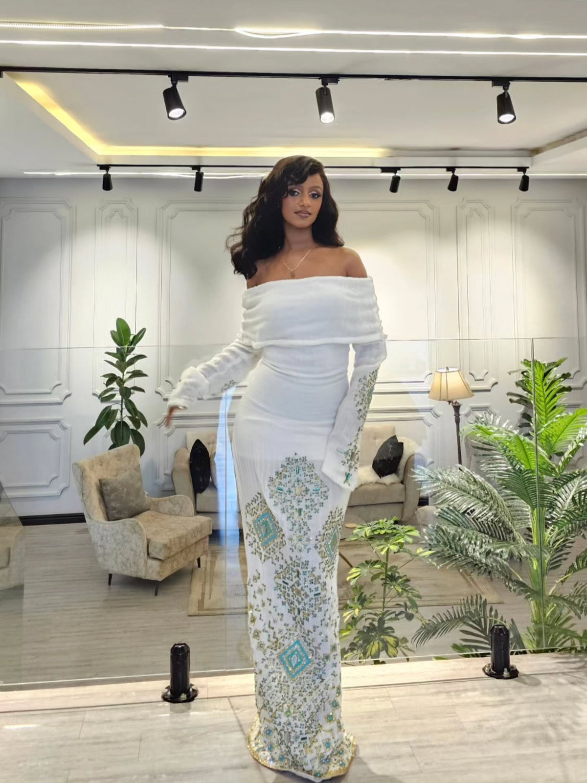 A woman models a white off-the-shoulder maxi Habesha Wedding Dress with long sleeves and intricate teal and gold floral embroidery down the skirt, standing in a modern, well-lit boutique showroom.