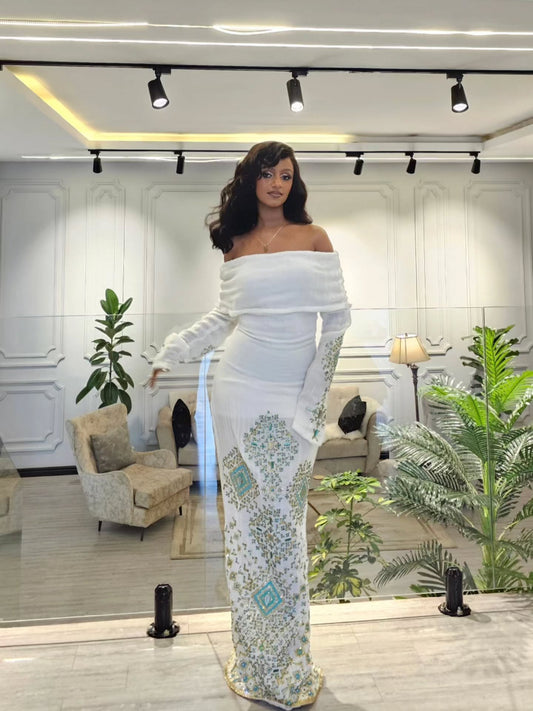 A woman models a white off-the-shoulder maxi Habesha Wedding Dress with long sleeves and intricate teal and gold floral embroidery down the skirt, standing in a modern, well-lit boutique showroom.