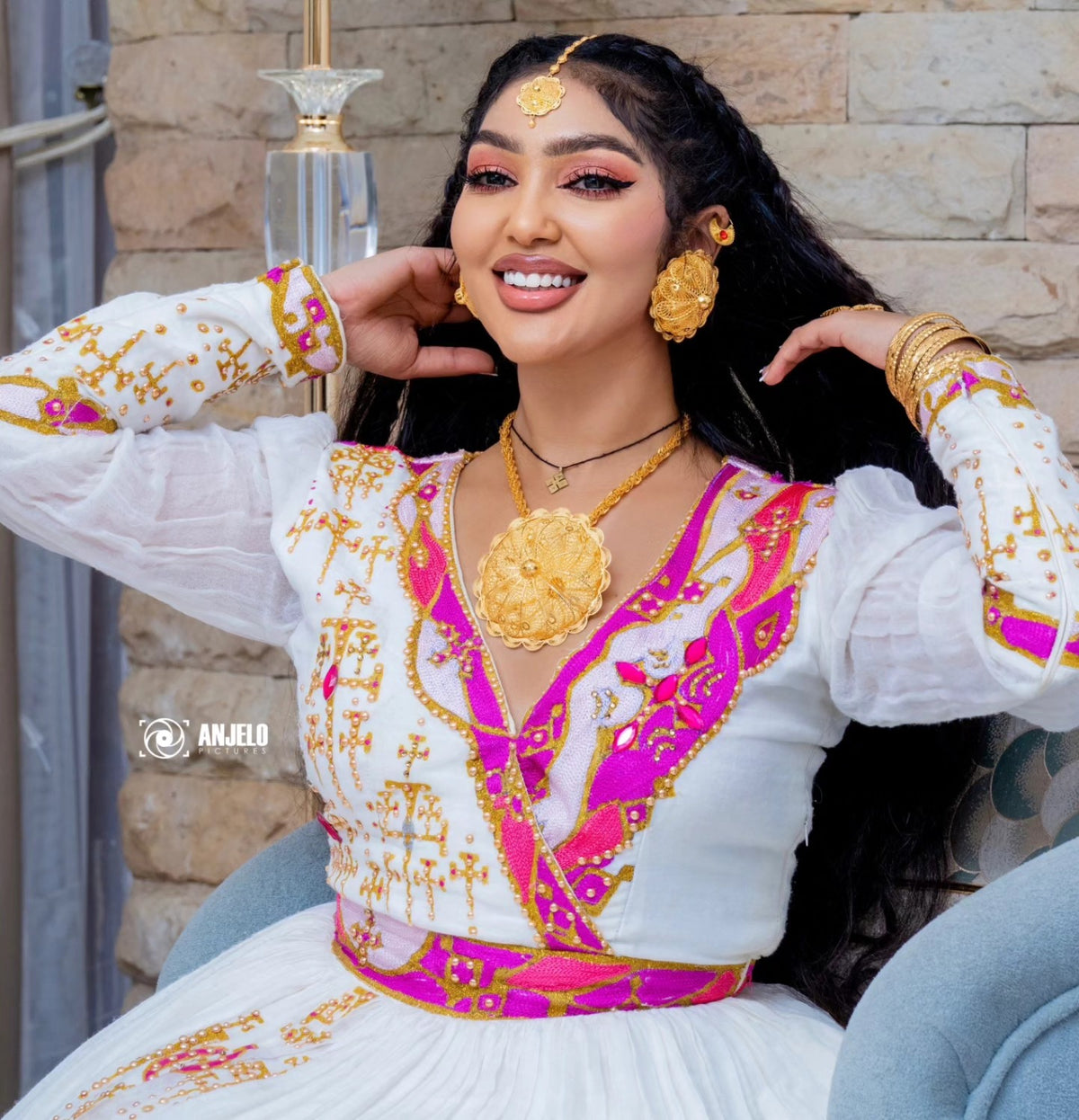 A woman wearing a traditional white embroidered Habesha wedding dress with bright pink and gold patterns, styled with gold jewelry, seated in a blue chair against a stone wall background.