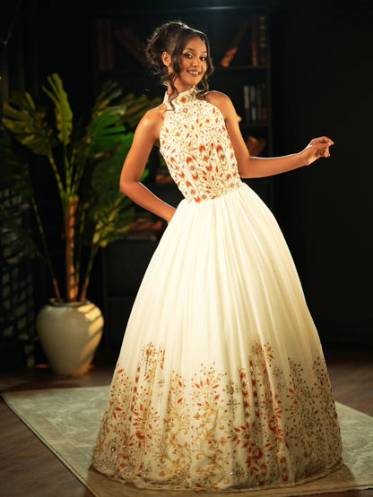 A woman models a formal ball Habesha Wedding Dress  with a high-neck bodice, cream tulle skirt, and orange floral embroidery, set against a dark indoor background with a potted plant.