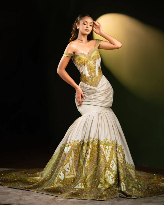 A model wears an elegant off-the-shoulder mermaid Habesha Wedding Dress  in off-white with extensive gold and olive-green sequined geometric patterns, set against a dark stage background with a spotlight.