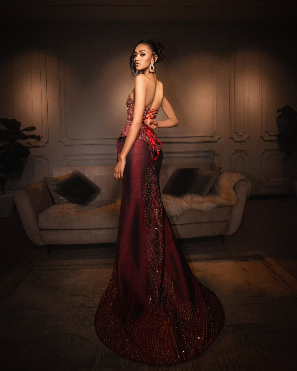 A woman stands in a dimly lit interior wearing a floor-length evening Habesha Wedding Dress. The dress is deep burgundy with ornate beaded and sequined detailing and a red sash at the waist.