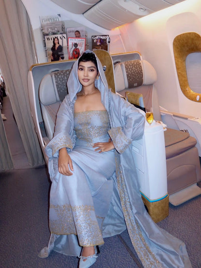 A woman wearing a light blue traditional Habesha Wedding Dress  with gold embroidery sits in an airplane seat. The dress has a matching head covering and a long draped train, with ornate gold detailing along the borders.