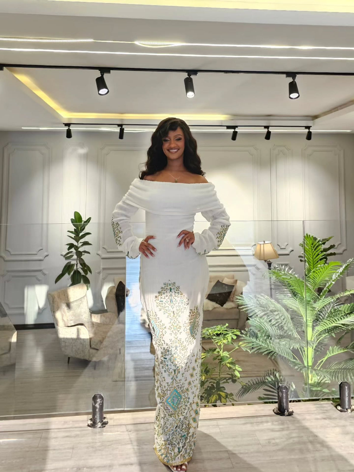 A woman stands indoors wearing a fitted off-the-shoulder white Habesha Wedding Dress with long sleeves and a heavily embellished multicolor floral pattern down the skirt and on the cuffs. The setting appears to be a modern showroom or lounge with track lighting, glass panels, plants, and upholstered seating in the background.