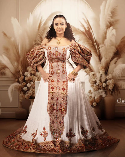 A woman stands indoors wearing an ornate off-shoulder traditional Habesha Wedding Dress  with a white skirt, heavily embroidered central panel, and voluminous brown patterned sleeves. The setting includes pampas grass decor and a warm beige interior backdrop.