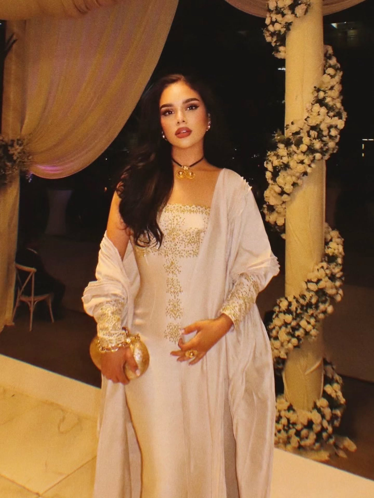 A woman wearing a white off-the-shoulder traditional Gold Habesha Wedding Dress  with gold embroidery and a matching long cardigan, standing in front of a decorated event backdrop with draped curtains and floral garlands.