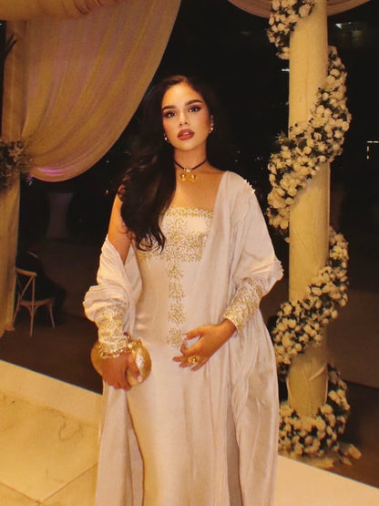 A woman wearing a white off-the-shoulder traditional Gold Habesha Wedding Dress  with gold embroidery and a matching long cardigan, standing in front of a decorated event backdrop with draped curtains and floral garlands.