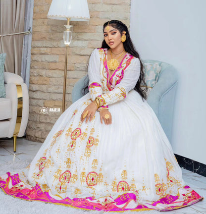 A woman seated in a light blue armchair wearing a white traditional Habesha wedding dress with bright pink and gold embroidery. The dress has long sleeves, a V-neckline, and a full skirt with ornate gold motifs and a wide pink embroidered border.