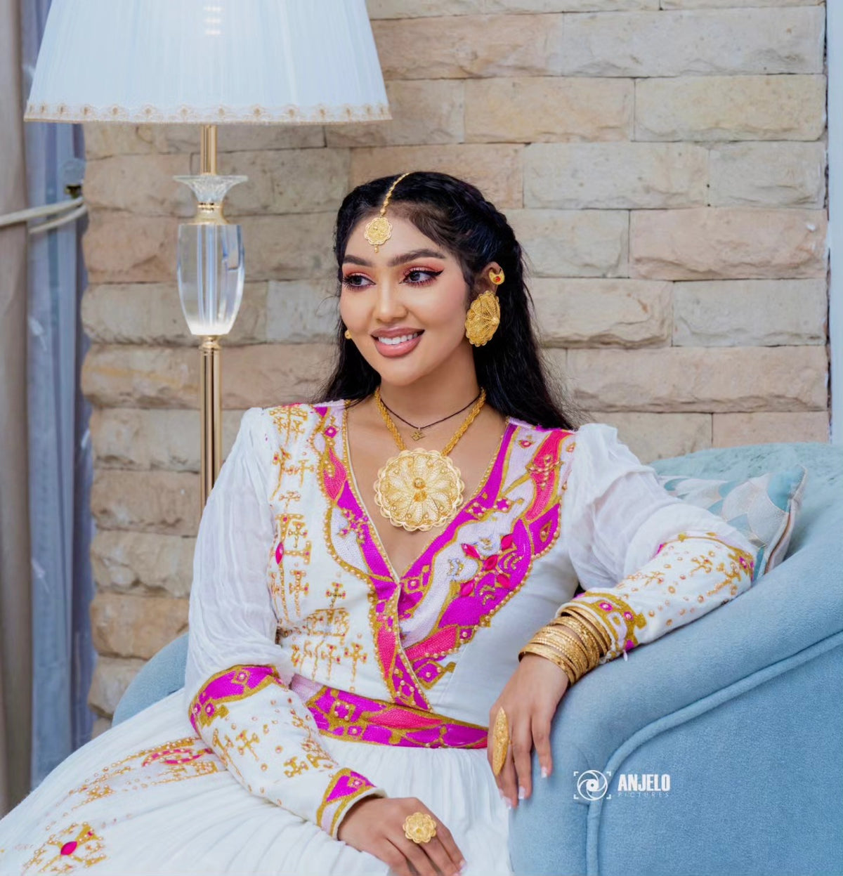 A woman wearing a white traditional Habesha wedding dress with bright pink and gold embroidery, seated on a light blue sofa against a beige stone wall. She is wearing extensive gold jewelry, including a large pendant, earrings, rings, and bangles.