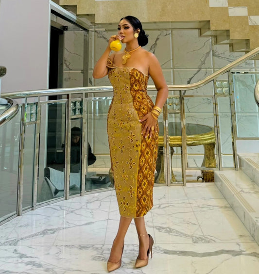 A woman stands in a modern marble lobby wearing a strapless golden-yellow habesha Wedding dress with a brown geometric pattern and nude heels, holding a yellow drink.