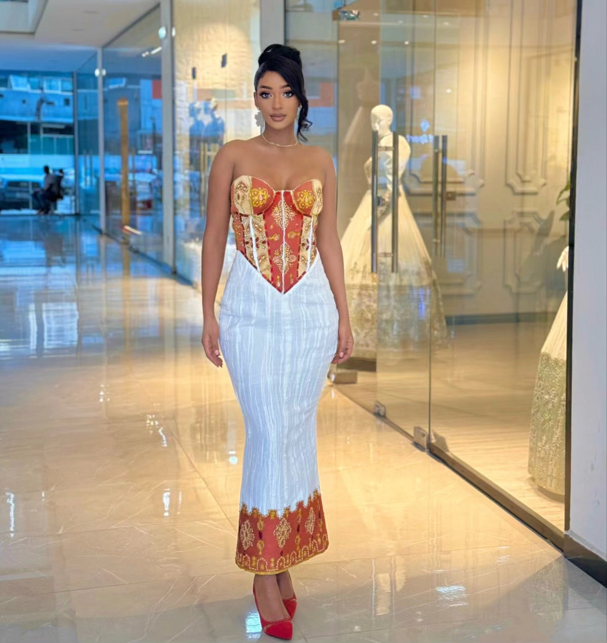 A woman models a strapless mermaid-style Habesha wedding dress with a red and gold corset-style bodice and a white pleated skirt with a red and gold patterned hem, standing in a modern glass-walled showroom.