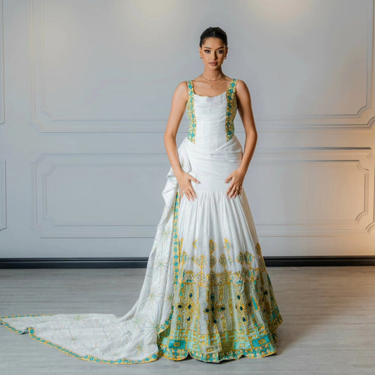 A woman stands indoors wearing a white sleeveless Habesha Wedding Dress with teal and gold ornate embroidery concentrated along the neckline, side panels, hem, and matching draped fabric on the floor.