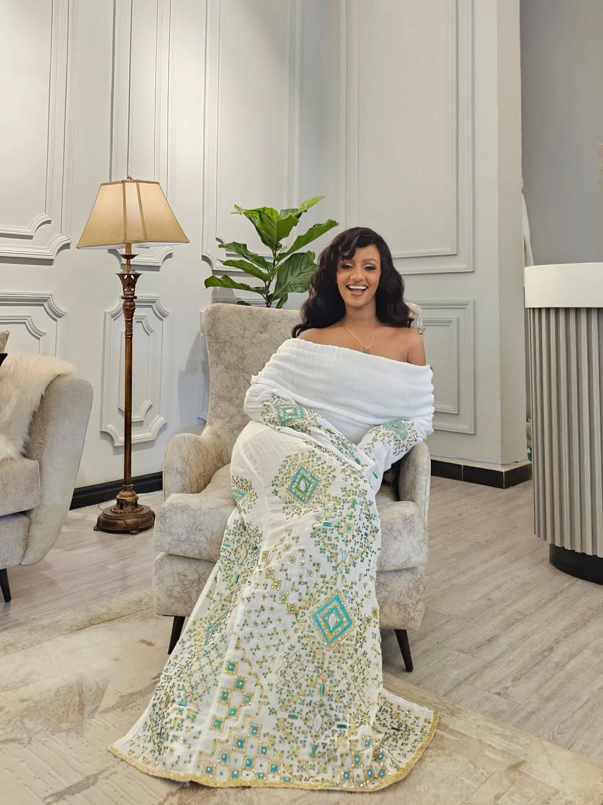 A woman sits in a light gray armchair wearing a white off-the-shoulder Habesha Wedding Dress with intricate teal and gold geometric embroidery, in a modern living room setting.
