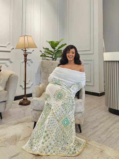 A woman sits in a light gray armchair wearing a white off-the-shoulder Habesha Wedding Dress with intricate teal and gold geometric embroidery, in a modern living room setting.
