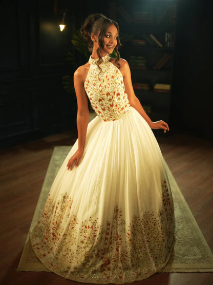 A young girl modeling a cream ball Habesha Wedding Dress with intricate floral embroidery in red, gold, and green, set against a dark, elegant indoor background.