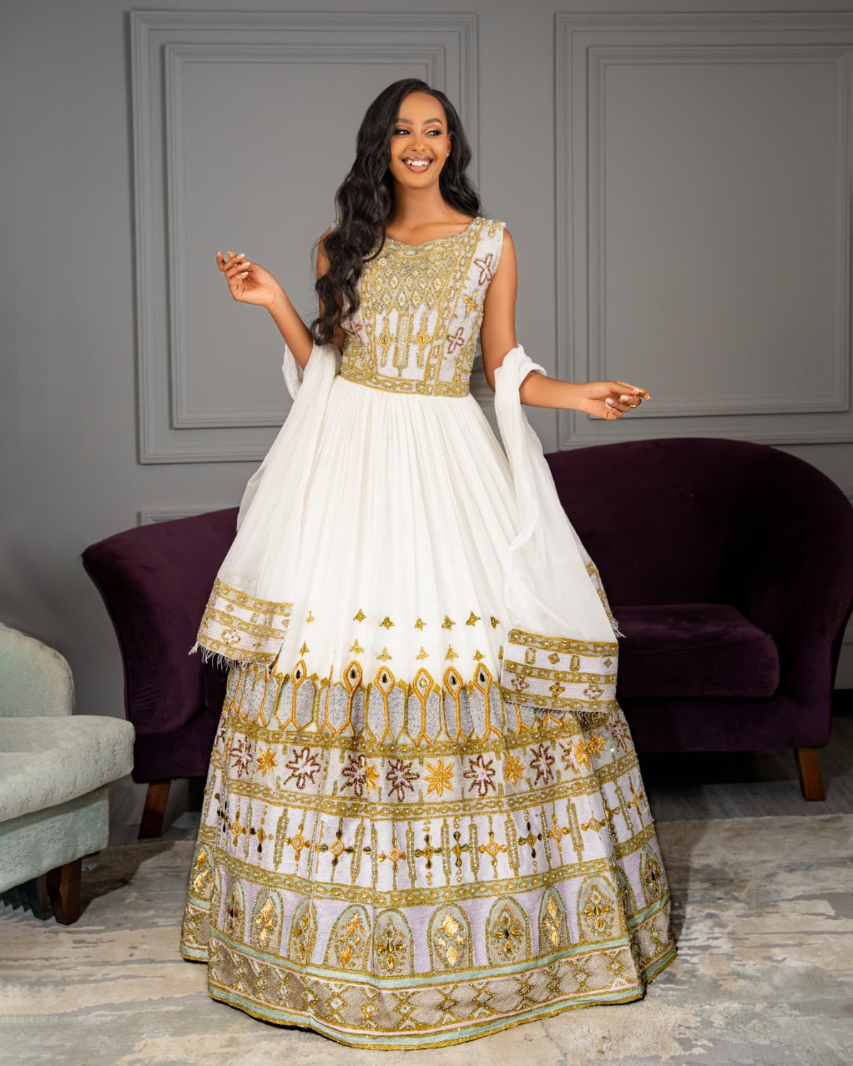 A woman modeling a white traditional Habesha Wedding Dress  with extensive gold and silver embroidery, standing in a room with gray paneled walls and purple velvet armchairs.