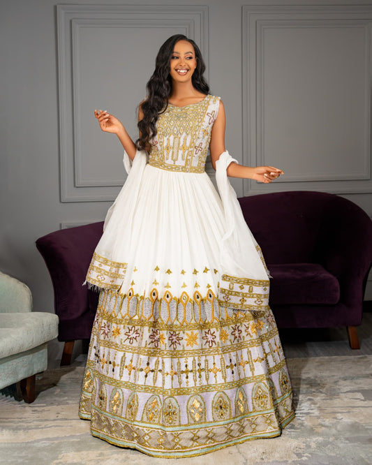 A woman modeling a white traditional Habesha Wedding Dress  with extensive gold and silver embroidery, standing in a room with gray paneled walls and purple velvet armchairs.