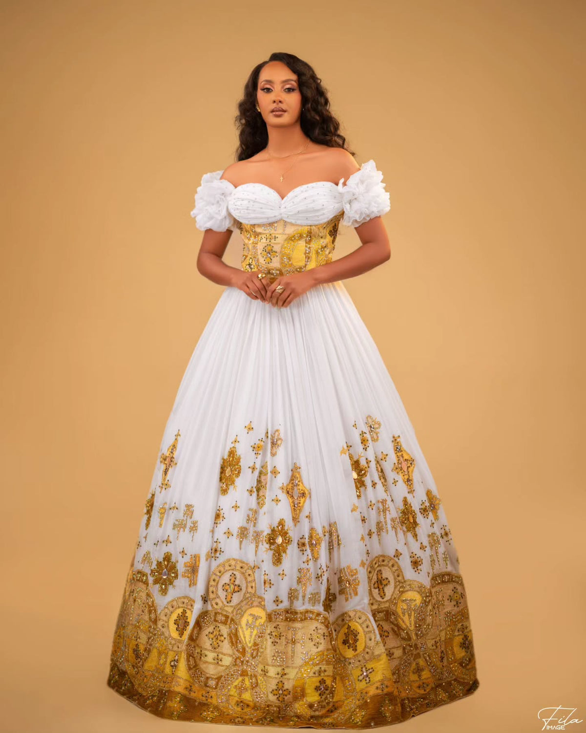 A woman modeling a grand off-the-shoulder Habesha Wedding Dress with a white sweetheart bodice, gold corset-style waist panel, and a voluminous white skirt decorated with gold floral and paisley motifs, set against a solid tan studio background.