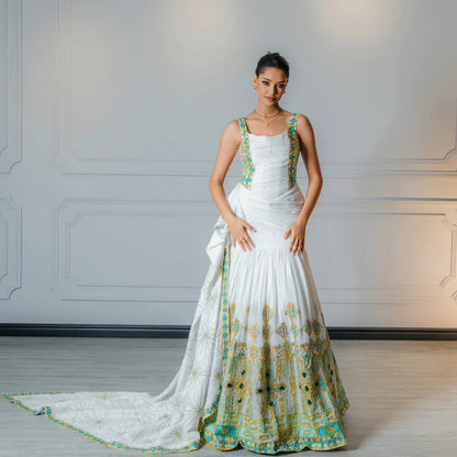 A woman models a white sleeveless traditional Habesha Wedding Dress with green and gold embroidery, standing against a light gray paneled wall. The dress features a draped dupatta with a matching embroidered border.