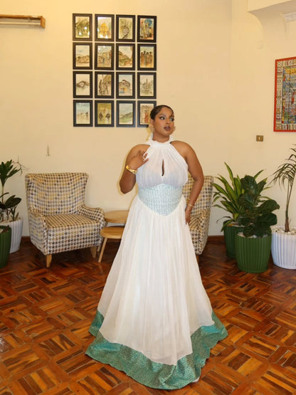 A woman modeling a white Habesha Wedding Dress with a light blue lace bodice and a teal patterned hem, standing in a furnished room with wooden floors, armchairs, and potted plants.