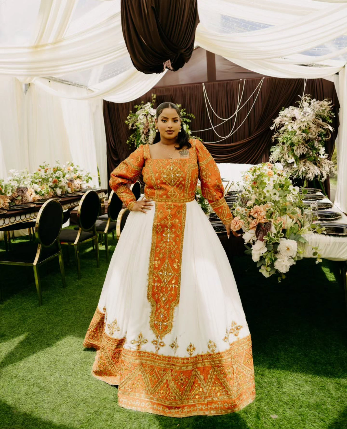 A woman stands in a decorated event tent wearing a traditional white Habesha Wedding Dress , a white center panel, and a prominent orange-gold embroidered border and matching embroidery on the bodice and sleeves.