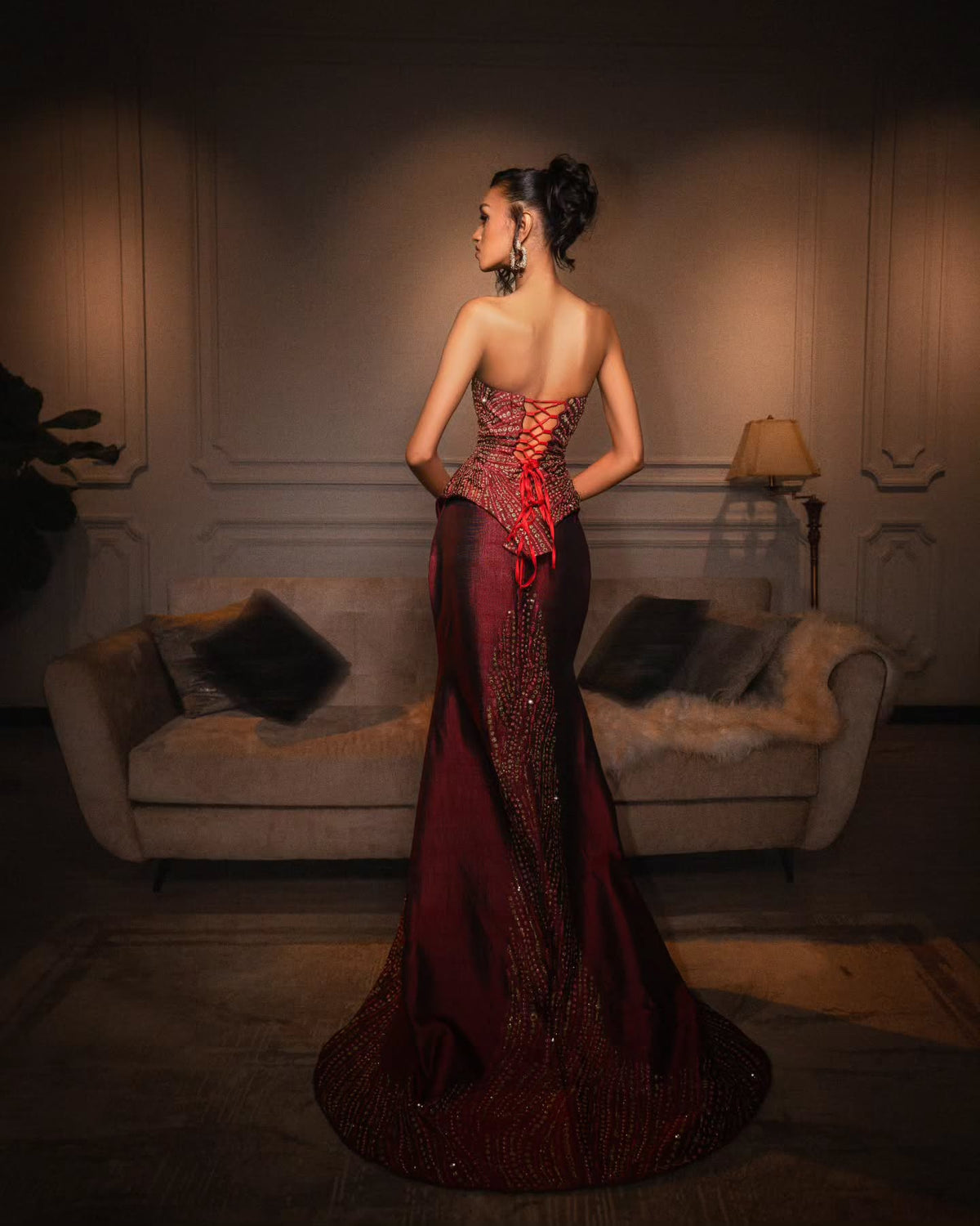 A woman stands with her back to the camera wearing a floor-length evening Habesha Wedding Dress in a dimly lit room with a sofa and lamp in the background. The dress is deep burgundy with a red lace-up corset-style back and shimmering embellished panels.