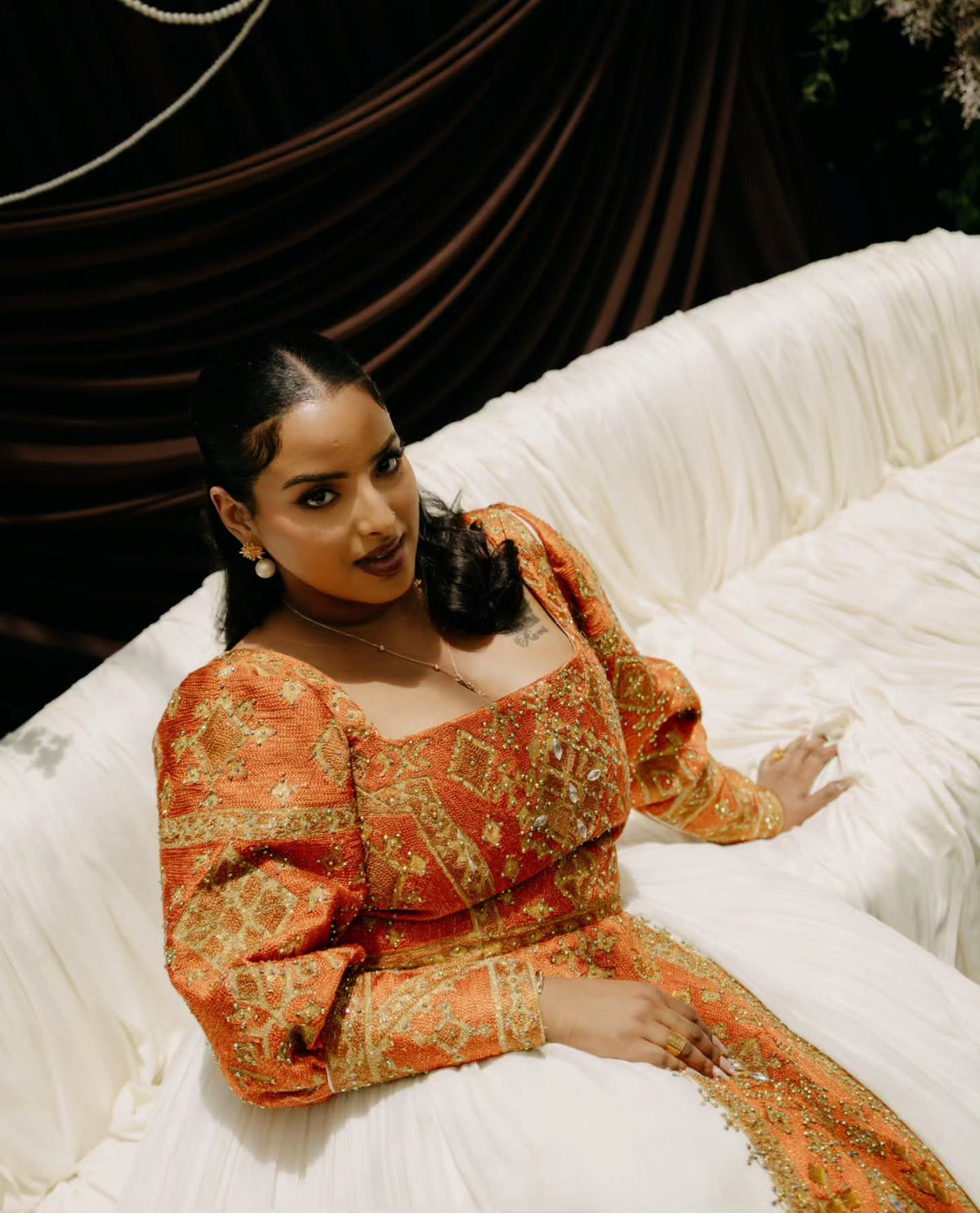 A woman reclining on a white draped sofa wearing a richly embroidered orange and gold traditional Habesha Wedding Dress  with puff sleeves and a square neckline.
