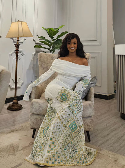 A woman wearing a white off-the-shoulder Habesha Wedding Dress with intricate teal and gold embroidery, seated in a plush armchair in a decorated room.