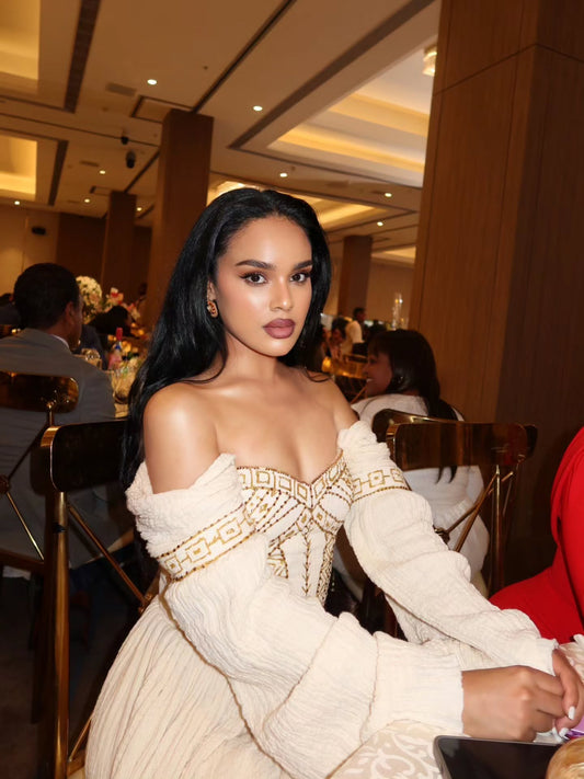 Woman in a white off-shoulder Habesha Wedding Dress with gold detailing at an indoor event.