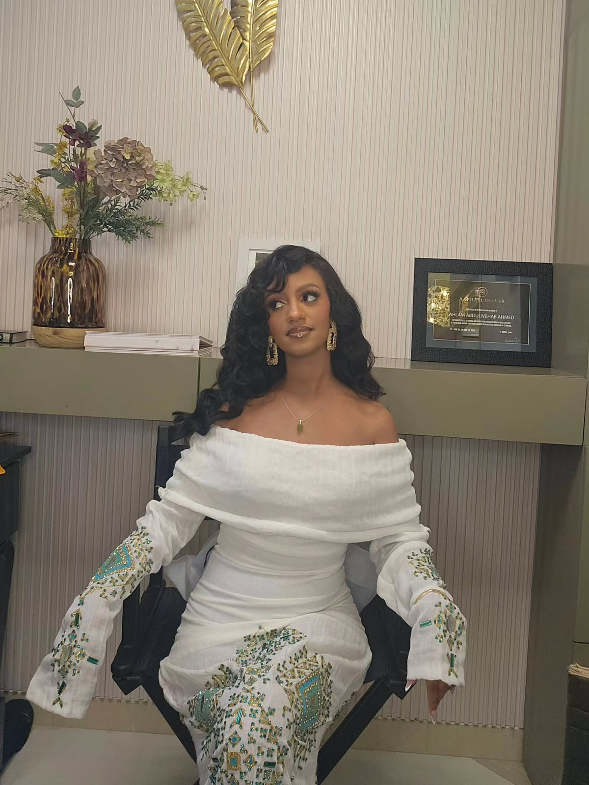 A woman seated in a salon chair wearing a white off-the-shoulder Habesha Wedding Dress with teal and green floral embroidery on the sleeves and skirt, in an indoor room with a shelf, framed certificate, and decorative wall piece.