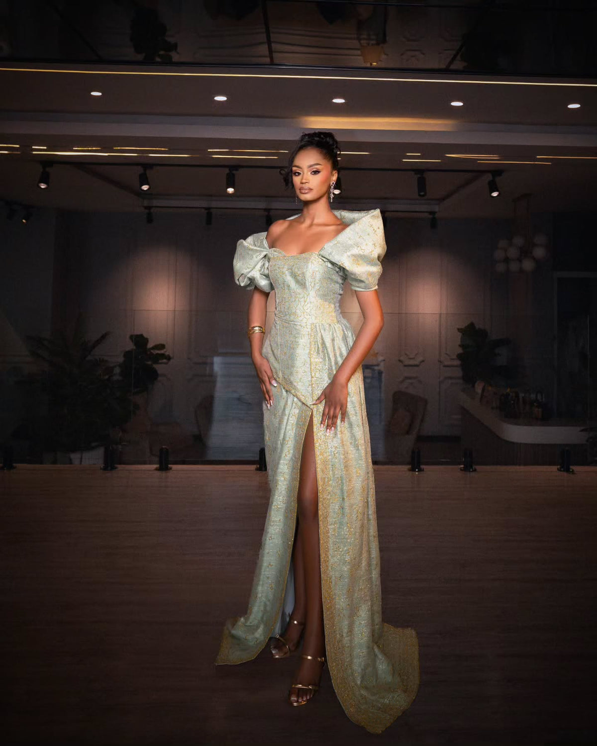 A model stands in a grand hall wearing a floor-length, off-the-shoulder Habesha Wedding Dress in a pale gold and silver patterned fabric with a high thigh slit.