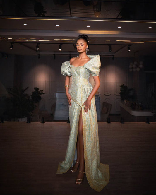 A model stands in a grand hall wearing a floor-length, off-the-shoulder Habesha Wedding Dress in a pale gold and silver patterned fabric with a high thigh slit.