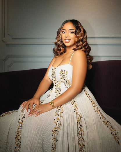 A woman wearing a white ball Gold Habesha Wedding Dress  with gold floral embroidery sits on a dark purple couch against a gray paneled wall.