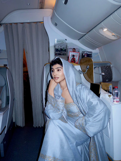 A woman wearing a light blue satin bridal Habesha Wedding Dress  with gold embroidery sits in an airplane first-class seat. The dress features a matching hood and a strapless neckline with wide sleeves.